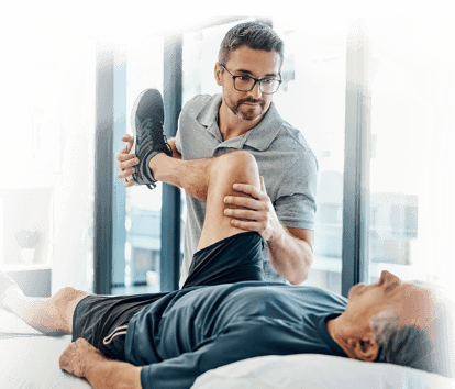 A physical therapist is helping an older man do leg stretches while lying on a bed.