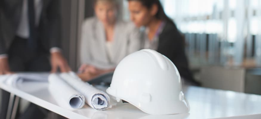 A close-up of a construction hat and two rolled up blueprints with business people in the background.